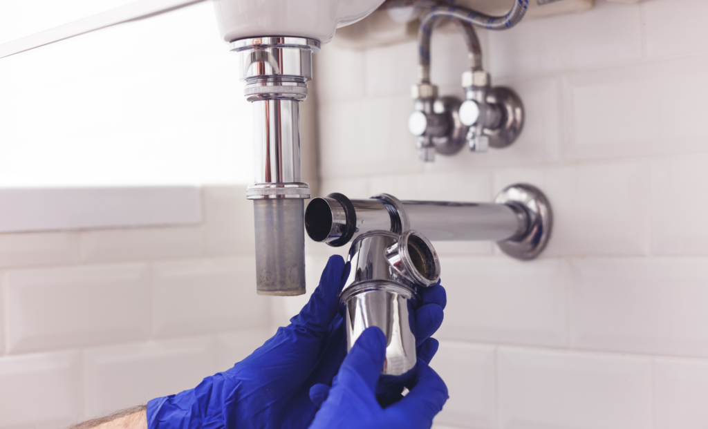 Gloved hands adjusting a chrome sink trap beneath a bathroom washbasin