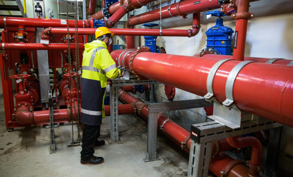 Worker in a high-visibility vest beside large red pipes in a mechanical room